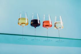 Four glasses of wine in a row on a glass shelf against a light blue wall