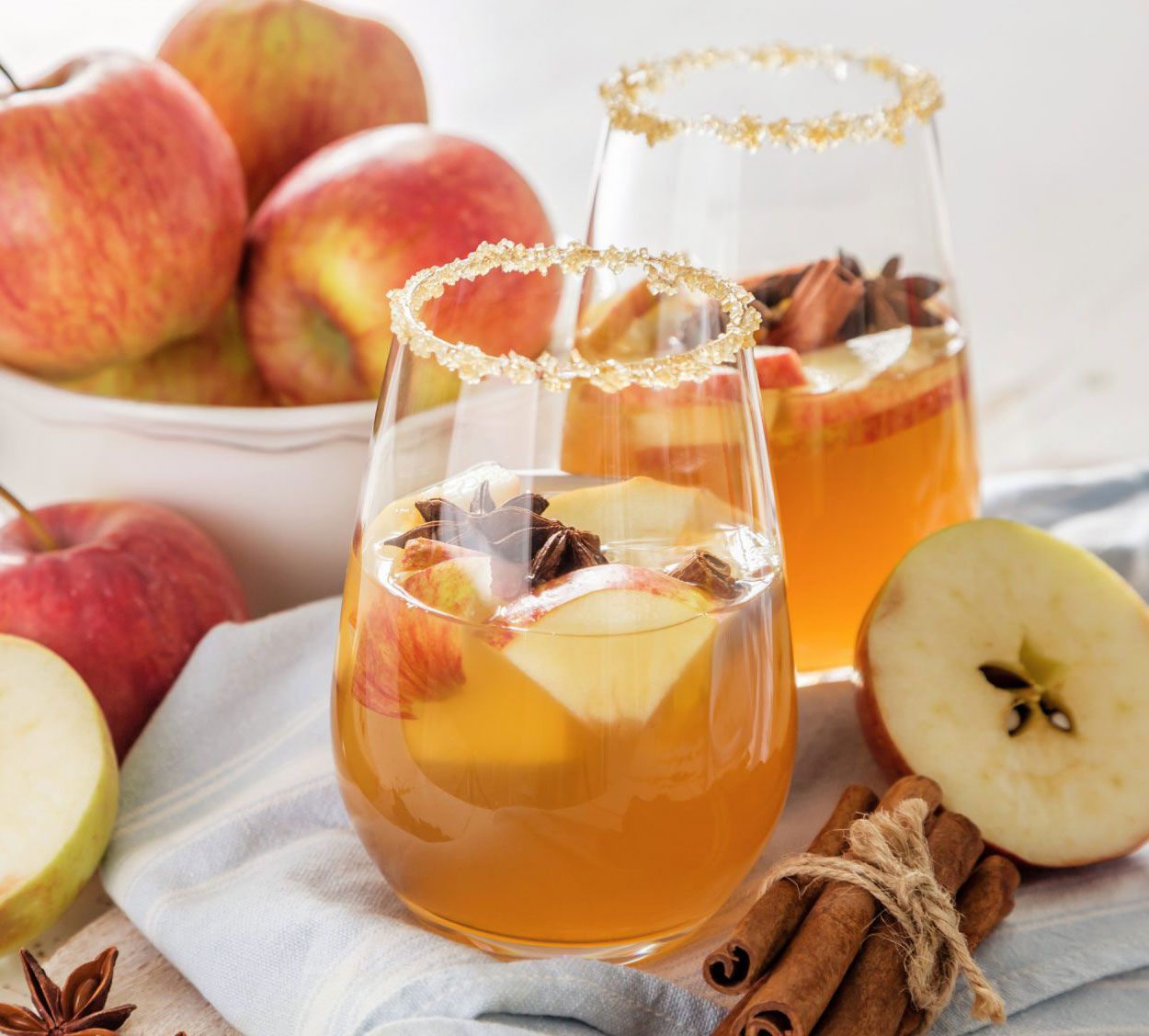 Apple Cinnamon-Spiced Cocktail Served in Two Glasses Garnished With Apples and Star Anise, Displayed on a Table With Fresh Apples and Cinnamon Sticks