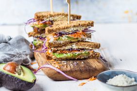 Multi-layer sandwich on cutting board with toothpick through the top, surrounded by half of an avocado with pit