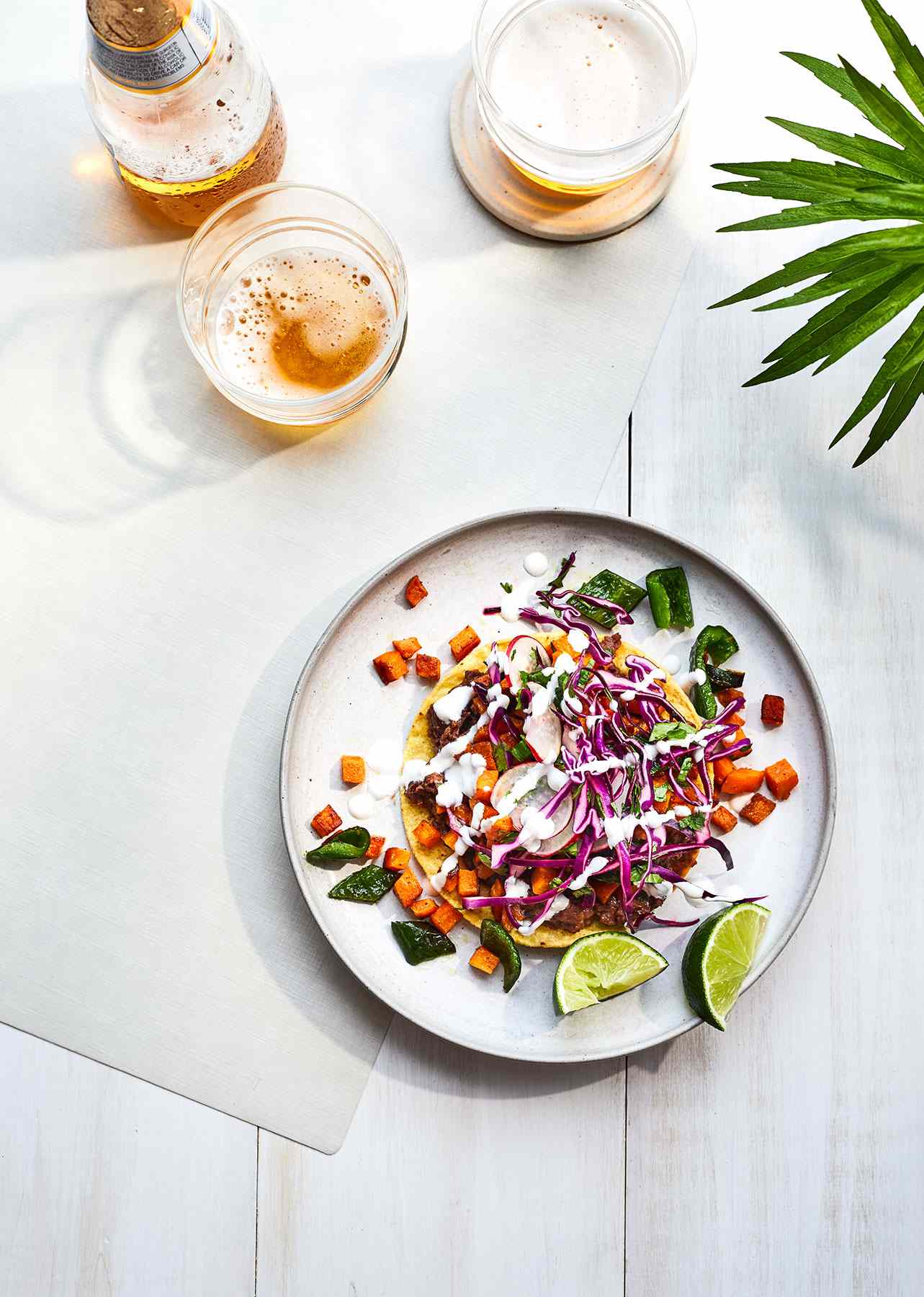 Black Bean Tostadas With Sweet Potato and Poblanos Served With Lime Wedges on a White Plate on a White Wooden Table, Surrounded by Beer Glasses