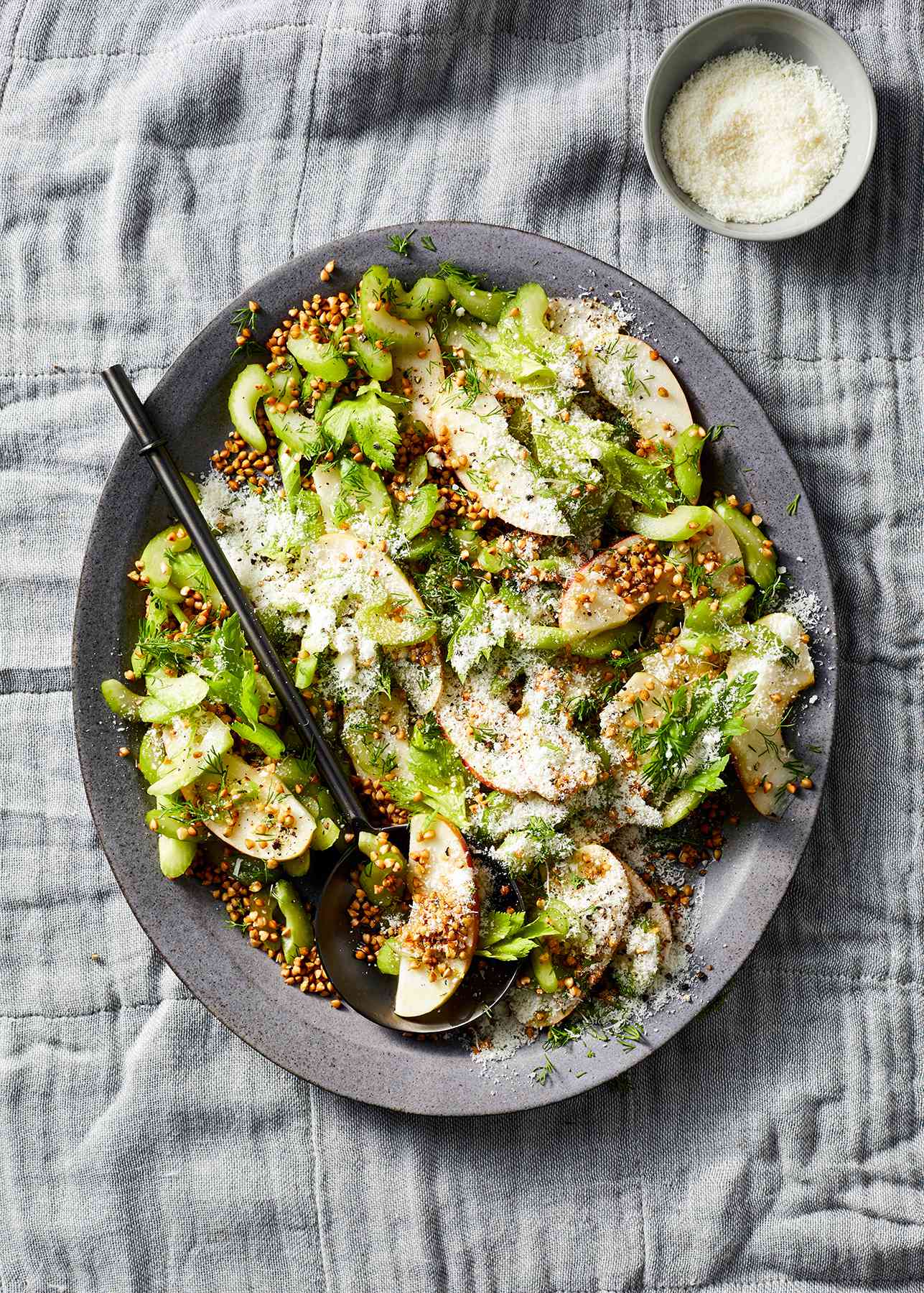 Celery and Apple Salad With Crispy Buckwheat Served on a Large Gray Plate With a Small Bowl of Grated Pecorino Romano Cheese on the Side