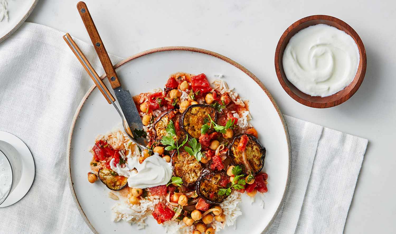 Eggplant-and-Tomato Masala With Chickpeas on a Bed of White Rice Served on a White Plate With Metal-and-Wood Utensils for Eating