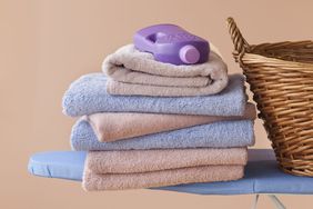 stack of five folded towels with a purple detergent bottle on top, sitting on a blue ironing board next to a wicker laundry basket