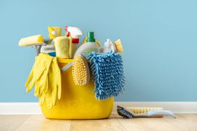 Kitchen cleaning brushes, liquids, sponges, and rubber gloves in a yellow bucket on the floor