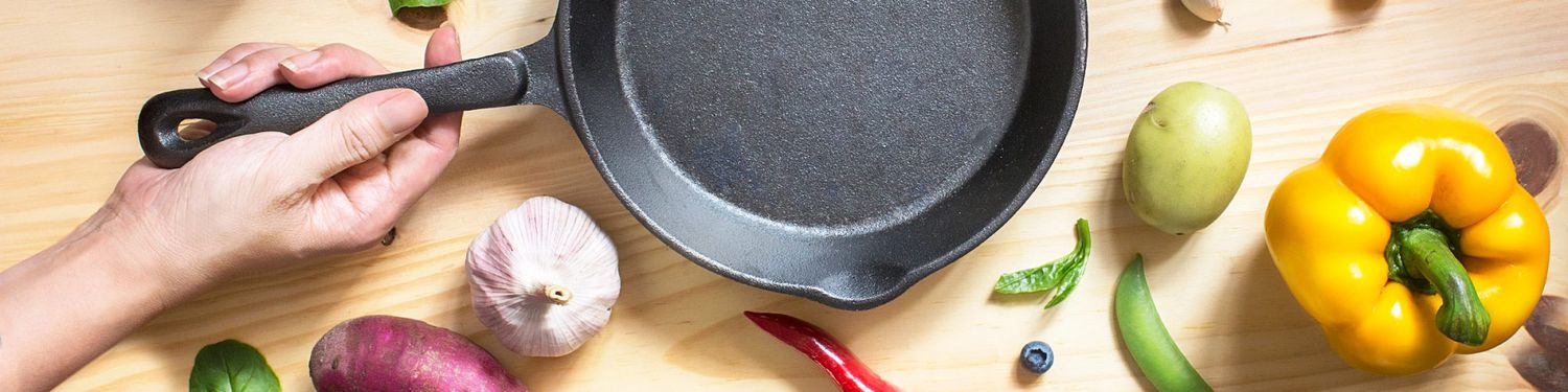 Person holding a cast-iron pan over a wooden surface surrounded by fresh vegetables ready for chopping