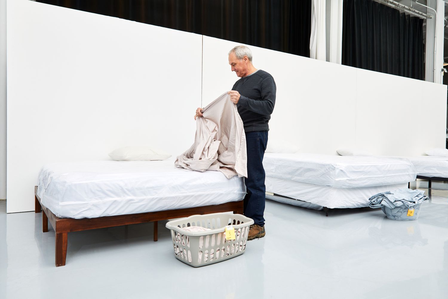 Man holding a sheet from the Parachute Sateen Sheet Set next to a bed and a laundry basket 