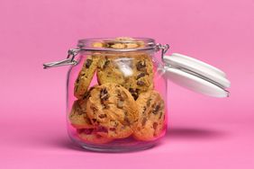 chocolate chip cookies in an open glass cookie jar against a pink background