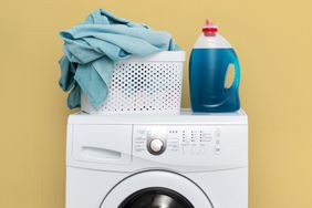 Concept of laundry process. White washing machine standing isolated inside bright light flat interior. Gel detergent soap with basket of lien on top of laundromat